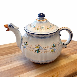 May include: A white ceramic teapot with a goose-shaped spout and handle. The teapot features a lid with a dark blue knob and is decorated with floral patterns in blue, green, and yellow. The teapot is placed on a wooden cutting board.