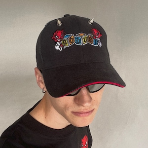 May include: Black baseball cap with red stitching and silver spikes. The cap features an embroidered design of two red cartoon devils with horns and a colorful block design that reads "DIALER".