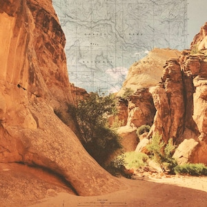 May include: A vintage map of Capitol Reef National Park in Utah, with the text "Capitol Reef Mesa National Park" printed on the map. The map is partially obscured by a view of a canyon with red rock formations and green vegetation.