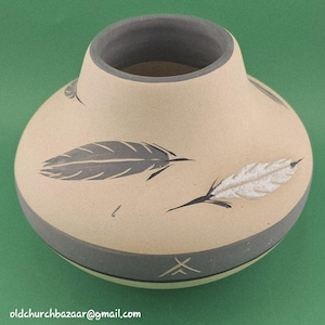 May include: A round, tan ceramic vase with a wide base and a narrow neck. The exterior features dark gray and white painted feather motifs. A solid dark gray band encircles the lower portion of the vase, and the interior rim is also dark gray.