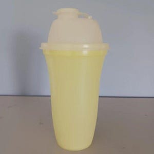May include: A pale yellow plastic shaker cup with a translucent, off-white lid. The container features a tapered cylindrical body and a secure, snap-on cap with a small spout opening at the top, designed for mixing liquids and ingredients.