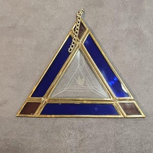 May include: A stained glass triangle with a gold-colored metal frame and chain. The triangle features blue and brown glass sections, with a clear glass center etched with a design. The chain is attached at the top.