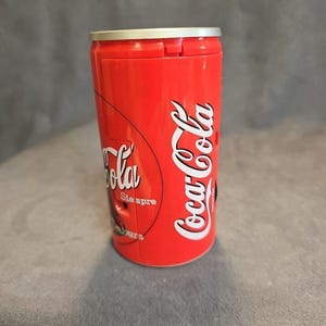 May include: A red Coca-Cola can-shaped container with white Coca-Cola branding. The container has a silver top and features the words "Siempre" and "ours" in white. The container is cylindrical and stands upright.