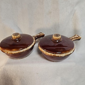 May include: Two brown ceramic soup bowls with lids. The bowls have a handle and a lid with a decorative knob. The rims and knobs have a cream-colored trim. The bowls are on a white surface.