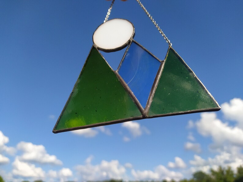 May include: A stained glass suncatcher featuring two green triangular mountain peaks, a central blue triangular peak, and a circular white sun positioned above the mountains. The glass pieces are joined by dark metal solder and a silver chain.