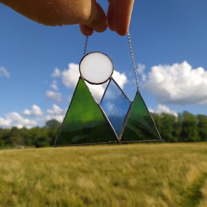May include: A stained glass suncatcher featuring a mountain landscape design with two dark green triangular peaks and one central blue triangular peak. A circular white glass piece sits above the blue peak, and the entire piece hangs from a silver chain.