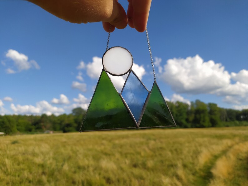 May include: A stained glass suncatcher featuring a mountain landscape design with two dark green triangular peaks and one central blue triangular peak. A circular white glass piece sits above the blue peak, and the entire piece hangs from a silver chain.