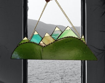 May include: A stained glass window hanging with a gold chain. The window depicts a green mountain range with blue and green peaks. The mountains are outlined in gold.