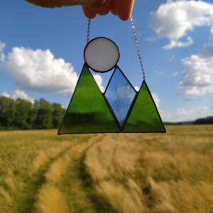 May include: A stained glass suncatcher featuring a mountain range design with two green triangular peaks and one central blue triangular peak. A white circular sun sits above the peaks. The piece is framed in black metal and hangs from a silver chain.