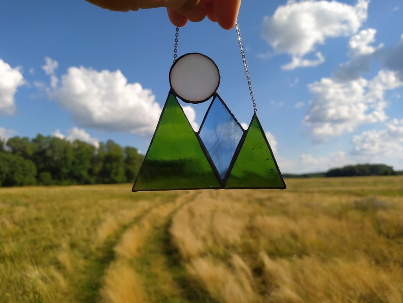 May include: A stained glass suncatcher featuring a mountain range design with two green triangular peaks and one central blue triangular peak. A white circular sun sits above the peaks. The piece is framed in black metal and hangs from a silver chain.