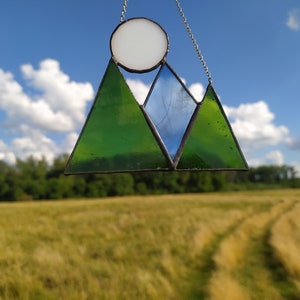 May include: A stained glass suncatcher featuring a mountain range design with two green triangular peaks and one central blue triangular peak. A circular white glass piece representing the sun is positioned above the mountains. The piece is held by a silver chain.