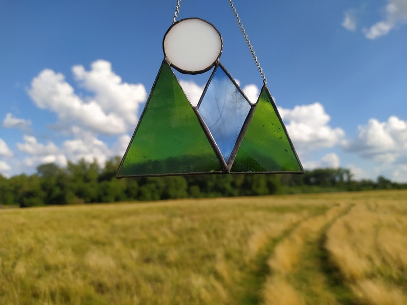 May include: A stained glass suncatcher featuring a mountain range design with two green triangular peaks and one central blue triangular peak. A circular white glass piece representing the sun is positioned above the mountains. The piece is held by a silver chain.