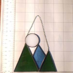 May include: A stained glass suncatcher featuring two dark green triangular mountains, a central blue triangular mountain, and a white circular moon. The piece is suspended by a silver chain and measures approximately 5.5 inches wide by 6 inches tall.