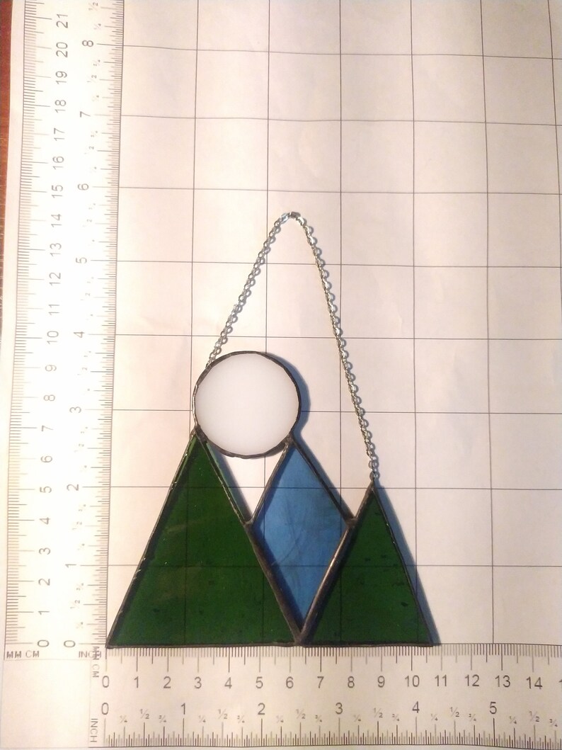 May include: A stained glass suncatcher featuring two dark green triangular mountains, a central blue triangular mountain, and a white circular moon. The piece is suspended by a silver chain and measures approximately 5.5 inches wide by 6 inches tall.