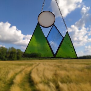 May include: A stained glass suncatcher featuring two green triangular mountain peaks and one blue triangular peak, topped with a circular white sun. The glass pieces are joined by dark metal solder lines and suspended by a thin silver-toned metal chain.