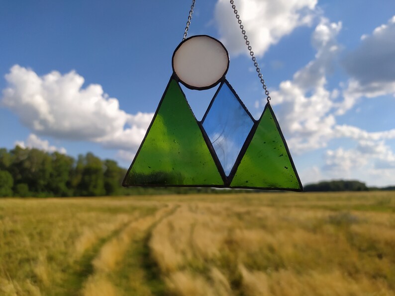 May include: A stained glass suncatcher featuring two green triangular mountain peaks and one blue triangular peak, topped with a circular white sun. The glass pieces are joined by dark metal solder lines and suspended by a thin silver-toned metal chain.