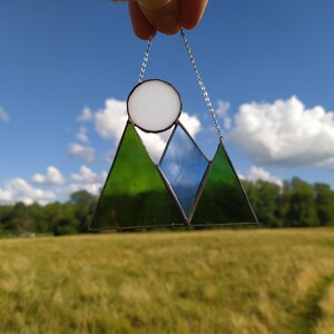 May include: A stained glass suncatcher featuring a mountain range design with two dark green triangular peaks and one central blue triangular peak. A circular white glass piece sits above the mountains, and the entire piece hangs from a silver chain.
