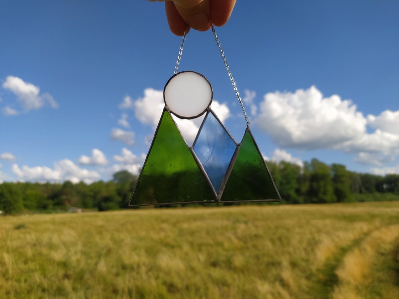 May include: A stained glass suncatcher featuring a mountain range design with two dark green triangular peaks and one central blue triangular peak. A circular white glass piece sits above the mountains, and the entire piece hangs from a silver chain.