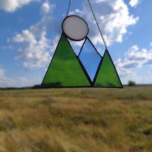 May include: A stained glass suncatcher featuring a mountain range design with two green triangular peaks and one blue triangular peak, topped by a white circular sun. The glass pieces are held together by dark metal solder and hang from a silver chain.