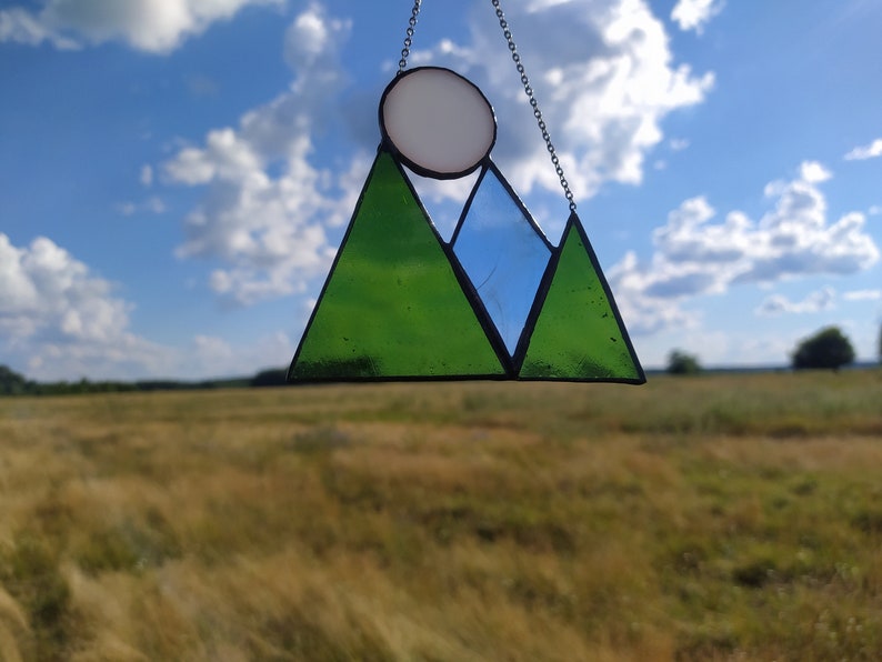 May include: A stained glass suncatcher featuring a mountain range design with two green triangular peaks and one blue triangular peak, topped by a white circular sun. The glass pieces are held together by dark metal solder and hang from a silver chain.