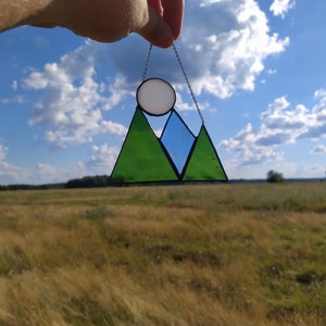 May include: A stained glass suncatcher featuring a mountain range design with two green triangular peaks and a central blue peak. A circular white glass piece sits above the mountains, representing a sun or moon, all held by a thin metal chain.