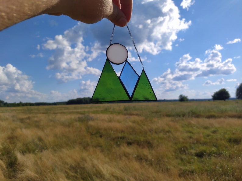 May include: A stained glass suncatcher featuring a mountain range design with two green triangular peaks and a central blue peak. A circular white glass piece sits above the mountains, representing a sun or moon, all held by a thin metal chain.