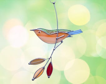 May include: A stained glass bird with a green, orange, and blue body perched on a branch with three leaves. The leaves are red, orange, and yellow.