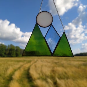 May include: A stained glass suncatcher featuring two green triangular mountain peaks, a central blue triangular peak, and a white circular sun above, all held together by dark metal solder lines and suspended by a thin silver-toned metal chain.