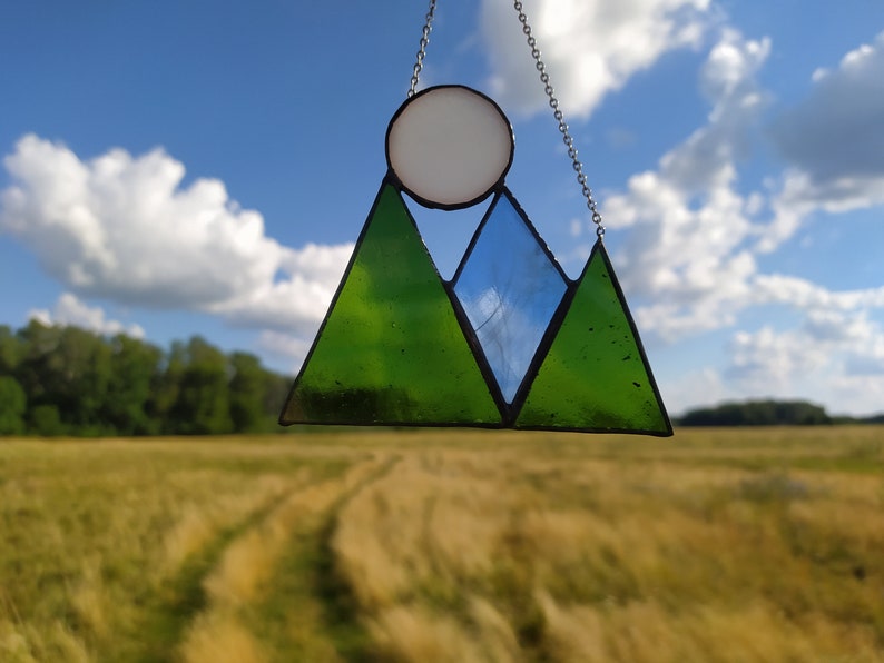 May include: A stained glass suncatcher featuring two green triangular mountain peaks, a central blue triangular peak, and a white circular sun above, all held together by dark metal solder lines and suspended by a thin silver-toned metal chain.