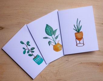 May include: Three watercolour illustrations of potted plants on white cards. The first card features a green plant with a blue pot. The second card features a green plant with a yellow pot. The third card features a green plant with a yellow pot on a metal stand.