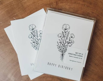 Puede incluir: Un juego de 12 tarjetas de felicitación con un diseño de ramo floral. Las tarjetas blancas tienen un dibujo lineal negro y las palabras "Happy Birthday". Se presentan en una funda transparente, sobre una superficie de madera.
