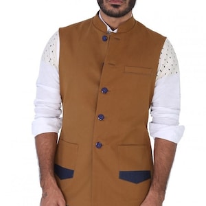 Op de afbeelding: Een bruin, mouwloos vest met een mandarijnkraag en blauwe knopen. Het vest heeft twee zakken aan de voorkant en een contrasterend blauw stoffen detail op de zakken.