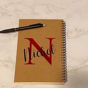 Puede incluir: Una libreta espiral marrón con una letra "N" roja y el nombre "Nichole" escrito en cursiva negra. Un bolígrafo negro está sobre la libreta.