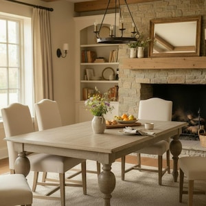 May include: A dining room scene featuring a light-colored wooden table with six upholstered chairs. A fireplace with a stone facade and a wooden mantel is in the background. A chandelier hangs above the table, and a window provides natural light.