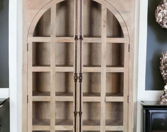 May include: A light-colored wooden cabinet with an arched top and glass-paned doors. The cabinet has multiple shelves and dark metal hardware. It is a tall, vertical piece of furniture, likely for storage or display.