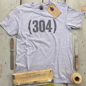 May include: Gray t-shirt with black text that reads "(304)". The shirt is laying flat on a wooden surface with a screen printing frame, a putty knife, and a ball of twine.