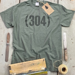 May include: A green t-shirt with the text (304) printed on the front. The t-shirt is laying on a wooden surface with a screen printing frame, a paint scraper, and a ball of twine.
