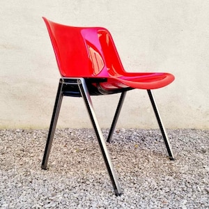 May include: A red plastic chair with a black metal frame. The chair is sitting on a gravel surface.