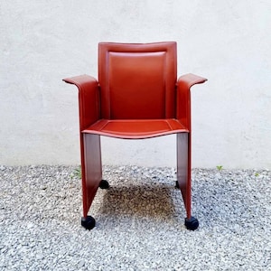 May include: A red leather office chair with a square frame and wheels. The chair is in front of a white wall.