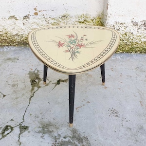 Puede incluir: Una mesa auxiliar de forma triangular con un diseño floral en la parte superior. La mesa tiene tres patas negras con puntas doradas.