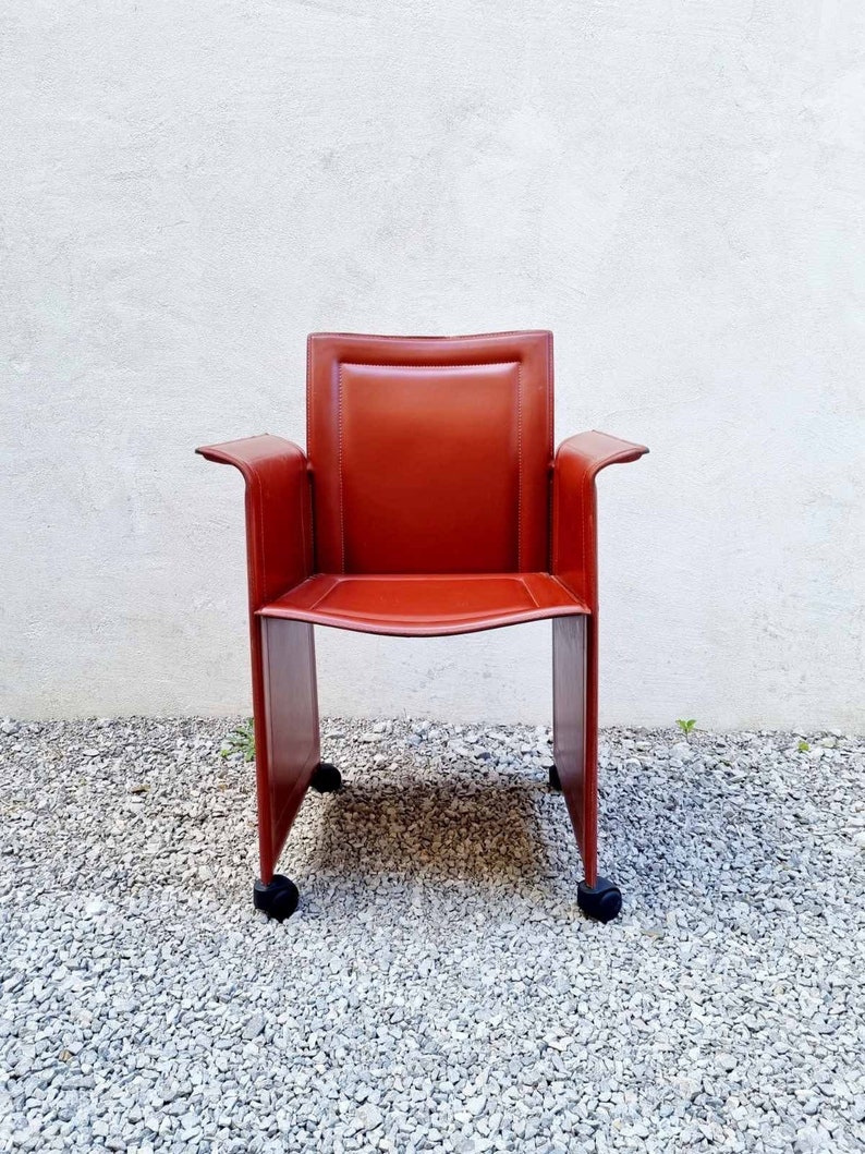 May include: A red leather office chair with a square frame and wheels. The chair is in front of a white wall.