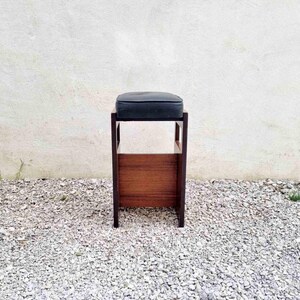 Puede incluir: Un taburete de madera con un coj&iacute;n oscuro. El taburete tiene un asiento rectangular y un panel inferior de madera. Las patas son oscuras y el taburete est&aacute; colocado contra una pared blanca y grava.