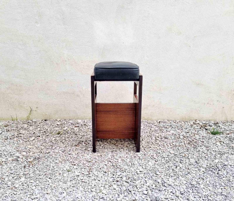 Puede incluir: Un taburete de madera con un coj&iacute;n oscuro. El taburete tiene un asiento rectangular y un panel inferior de madera. Las patas son oscuras y el taburete est&aacute; colocado contra una pared blanca y grava.