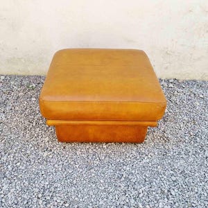 May include: A square, mid-century modern ottoman in a warm, caramel-colored leather. The ottoman has a cushioned top and a sturdy, rectangular base. It is placed on a bed of small, gray gravel.