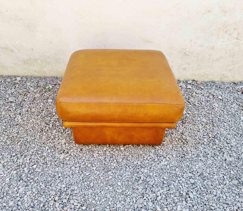 May include: A square, mid-century modern ottoman in a warm, caramel-colored leather. The ottoman has a cushioned top and a sturdy, rectangular base. It is placed on a bed of small, gray gravel.
