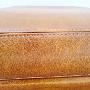 May include: Close-up of a section of a tan leather item. The leather has a textured surface with visible grain and subtle color variations. The item is stitched with a contrasting thread, and the overall appearance suggests a high-quality product.