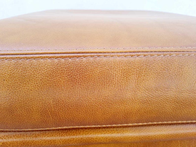 May include: Close-up of a section of a tan leather item. The leather has a textured surface with visible grain and subtle color variations. The item is stitched with a contrasting thread, and the overall appearance suggests a high-quality product.