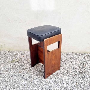 Puede incluir: Un taburete de madera vintage con un asiento acolchado negro. El taburete tiene un asiento rectangular y un marco de madera oscura. El marco tiene un dise&ntilde;o recortado en los lados. El taburete est&aacute; colocado sobre una superficie de grava.