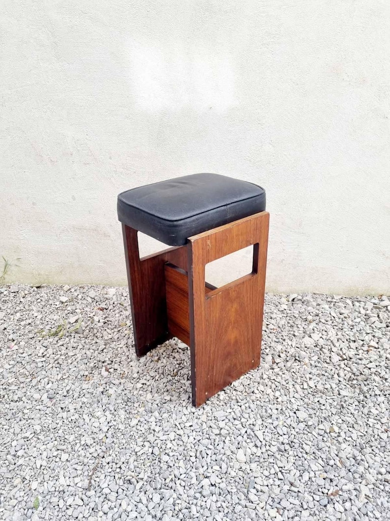 Puede incluir: Un taburete de madera vintage con un asiento acolchado negro. El taburete tiene un asiento rectangular y un marco de madera oscura. El marco tiene un dise&ntilde;o recortado en los lados. El taburete est&aacute; colocado sobre una superficie de grava.