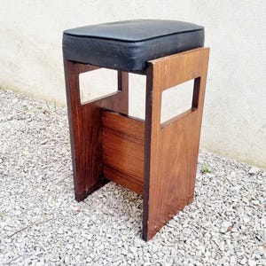Puede incluir: Taburete de madera vintage con asiento acolchado negro. El taburete tiene un asiento rectangular y un dise&ntilde;o de marco abierto con detalles recortados. La madera tiene un tono marr&oacute;n rojizo c&aacute;lido, sobre grava.
